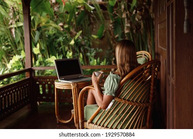 Young woman reading document and working on laptop computer sitting at table on wooden balcony of country house and drink tea