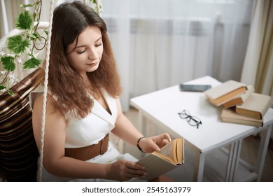 A young woman reading intently while sitting on a cozy indoor swing beside a table with books and glasses in a sunlit room - Powered by Shutterstock