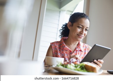 A young woman reading from the screen of a digital tablet, seated at a table. Coffee and a sandwich. - Shutterstock ID 1664716828