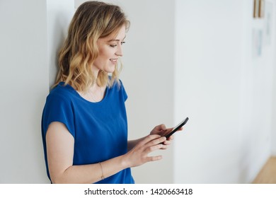 Young Woman Reading A Text Message On A Mobile Phone As She Leans Against An Interior White Wall At Home With Copy Space