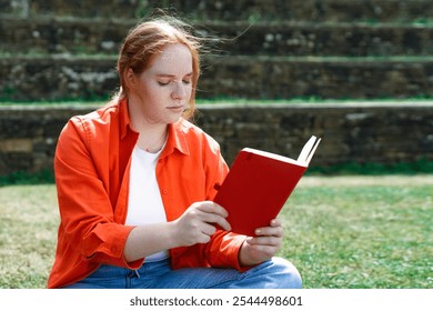 A young woman reads a book while seated on grass in a tranquil park setting during a sunny afternoon - Powered by Shutterstock