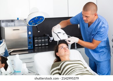 Young Woman Receiving Facial Light Therapy In Aesthetic Medicine Office. Cosmetologist Putting LED Mask On Face