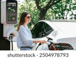 Young woman recharging battery for electric car during road trip travel EV car in natural forest or national park. Eco friendly travel during vacation and holiday. Exalt