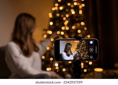 Young woman recording holiday video with decorated Christmas tree in background, cozy festive atmosphere at home - Powered by Shutterstock