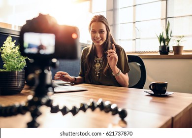 Young woman recording video for her vlog on a digital camera mounted on flexible tripod. Smiling woman sitting at her desk working on a laptop computer.