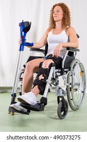 Young Woman Recovering From Knee Surgery Sitting In A Wheelchair With Her Leg In A Brace Holding Crutches Looking Aside With A Serious Expression