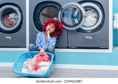 A Young Woman With Red Afro Hair Squatting Holding A Basket With Clothes In A Blue Automatic Laundromat