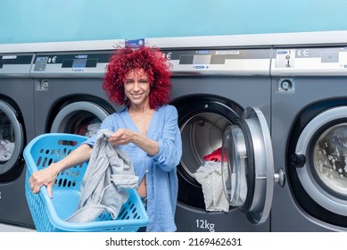 A Young Woman With Red Afro Hair Putting Dirty Clothes From The Laundry Basket Into The Washing Machine In A Blue Automatic Laundry Room.