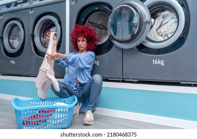 A Young Woman With Red Afro Hair Putting Dirty Clothes From The Laundry Basket Into The Washing Machine In A Blue Automatic Laundry Room.