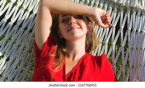 Young woman in red beach dress relaxing in comfortable hammock in a green garden on sunset. Summer luxury vacation, sunbathing, holidays concept. Thailand, Asia travel. Slow motion, close up