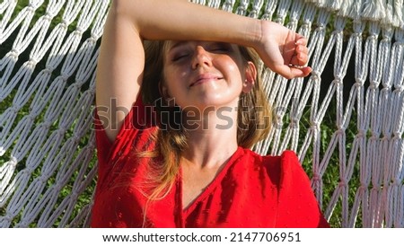 Young woman in red beach dress relaxing in comfortable hammock in a green garden on sunset. Summer luxury vacation, sunbathing, holidays concept. Thailand, Asia travel. Slow motion, close up