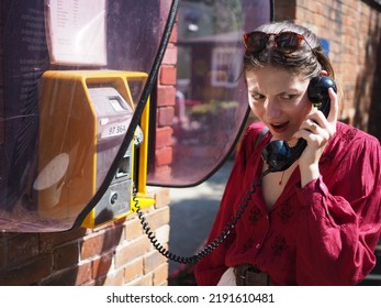 Young Woman In Red Blouse Making A Call Using A Public Pay Phone With Shocked Expression.