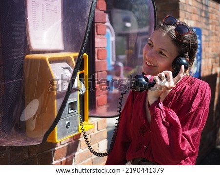 Young woman in red blouse using public telephone she is smiling and happy in conversation. Blurred background.