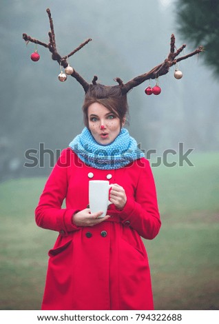 A young woman in the red coat with a cup of coffee in the character of a christmas deer in the foggy park. Christmas cheer. Christmas mood.