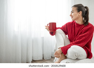 A young woman with a red cup of hot drink in a cozy red sweater is resting on the couch at home copy space.
