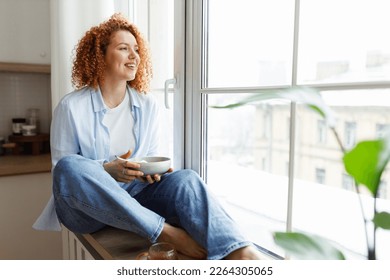 Young woman with red curly hair in jeans and shirt sitting on window sill looking through big window on city holding bowl of breakfast in hands, smiling noticing something funny outside - Powered by Shutterstock