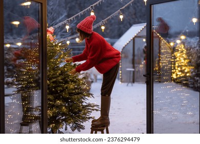 Young woman in red decorates lush Christmas tree with festive ballls and garland at backyard of her house on snow fall, preparing for a winter holidays
