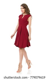 Young Woman In Red Dress And High Heels Is Walking, Looking Away And Smiling. Front Side View. Full Length Studio Shot Isolated On White.