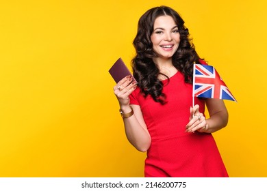 Young woman in red dress showing small flag UK and passport on yellow background in studio. Study abroad concept. International student exchange program. Learning British English with native speaker.
