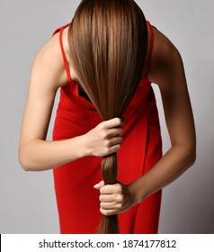Young woman in red dress standing with head down and holding her strong healthy long silky straight hair in hands over grey wall background. Haircare beauty, wellness, hairstyle, straightening concept
