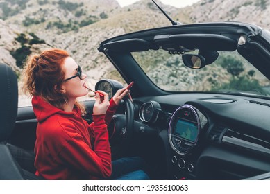 Young woman with red hair is applying lipstick looking at the mirrow in cabriolet with roof that folds down, summer vacation, staycation, enjoying life - Powered by Shutterstock