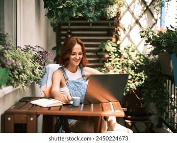 A young woman with red hair enjoys working on her laptop surrounded by lush potted plants on a sunny balcony - Powered by Shutterstock