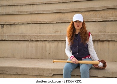Young woman with red hair, freckles, wearing white cap, jacket, baseball bat and glove, sitting on concrete bleachers. Concept sport, baseball, model, sports fashion, beauty.