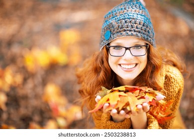 A young woman with red hair and glasses, wearing a knit hat and mustard-yellow sweater, smiles brightly while holding a handful of colorful autumn leaves outdoors on a sunny fall day.
 - Powered by Shutterstock