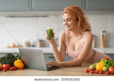 A young woman with red hair smiles as she drinks a green smoothie. She is seated at a kitchen counter, using a laptop while surrounded by colorful vegetables and fresh produce. - Powered by Shutterstock