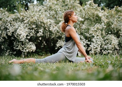 Young woman with red hair trains, doing exercises in yoga pose of a rider outdoors in the park.
