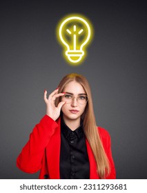 Young woman in red jacket with glowing bulb over head. Over gray background. - Shutterstock ID 2311293681