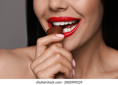 Young Woman With Red Lips Eating Heart Shaped Chocolate Candy On Grey Background, Closeup