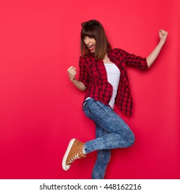 Young Woman In Red Lumberjack Shirt, Jeans And Brown Sneakers Standing On One Leg, Flexing Muscles, Looking Over The Shoulder And Shouting. Three Quarter Length Studio Shot On Yellow Background.