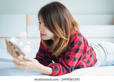 Young woman in red plaid shirt lying on bed, using smartphone with floating emoji reactions, expressing online interaction. - Powered by Shutterstock