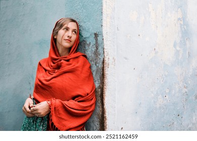 A young woman in a red shawl, leaning against a wall. The woman, wearing a red shawl, looks thoughtful. The red shawl and serene expression highlight the woman's peaceful demeanor. - Powered by Shutterstock