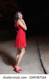 Young Woman In Red Sleeveless Dress Walking In Night Park. Attractive Long Haired Brunette In Red Sundress, Red Shoes With Small Red Bag In Her Hands Looking Over Shoulder At Camera