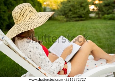 Young woman in a red swimsuit and a wide-brimmed hat is reading a book sitting on a chaise longue and relaxing. A woman enjoys life in the backyard with flowers and plants