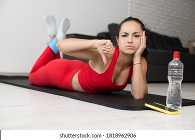 Young Woman In Red Tracksuit Doing Exercise Or Yoga At Home. Upset Unhappy Slim Sporty Model Lying On Mat And Holding Big Thumb Down. Dislike Or Negative Feedback About Training Or Workout.