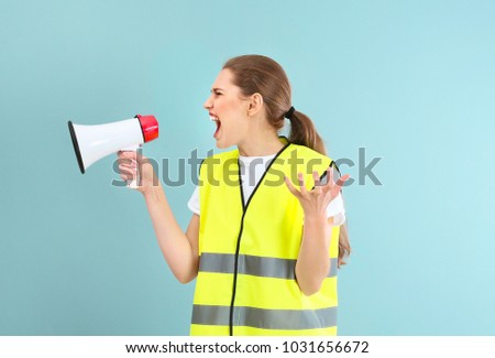 Young woman in reflective vest shouting into megaphone on color background
