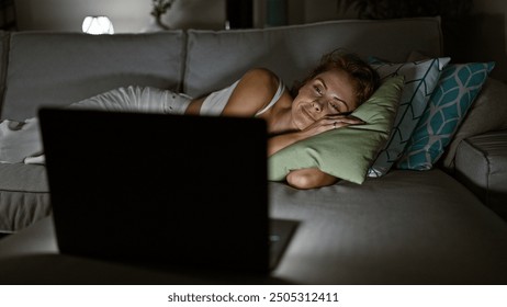 A young woman relaxes on a couch at home, watching a laptop in a cozy, dimly lit living room. - Powered by Shutterstock