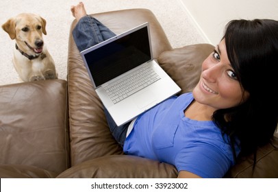 A Young Woman Relaxes On The Couch While On Her Laptop Computer With Her Dog At Her Side