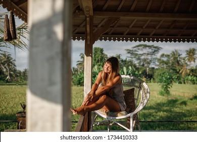 Young Woman Relaxing At The Beautiful Terrace While Sitting On The Chair With Green Rice Field Nature On Background. Luxury Hotel Home Living Woman Relax Enjoying Sofa Furniture Of Outdoor Patio