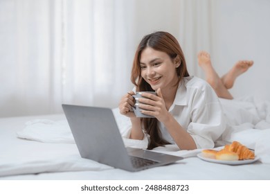 Young woman is relaxing in bed with a cup of coffee and pastries while working on her laptop, enjoying a leisurely and productive morning - Powered by Shutterstock