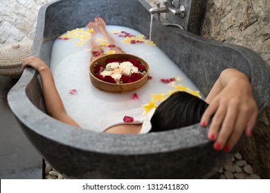 Young Woman Relaxing In Black Stone Bath With Tropical Flowers And Rose Petals. Skin Treatment, Luxury Spa Concept.