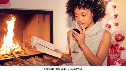 Young Woman Relaxing With A Book And Red Wine At Christmas Sitting In Front Of A Burning Fire Alongside A Decorated Tree