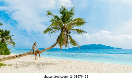 Young woman relaxing at a coconut palm tree on a white tropical beach at La Digue Seychelles Islands. Asian woman with hat sitting on a palm tree looking out over the ocean - Powered by Shutterstock