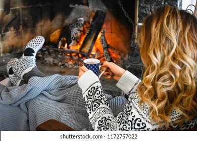 Young Woman Relaxing In Front Of Cozy Fireplace And Warming Up Her Feet In Woolen Socks In Country House. Woman Holding Mug Of Tea Or Coffee. Winter Or Autumn Vacation. Christmas Holidays Concept. 
