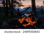 A young woman relaxing in a hammock near a campfire, looking at the Red Deer River at the Eagle Creek Campground in Yaha Tinda, Alberta, Canada.