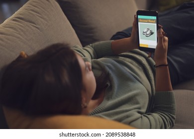 Young Woman Relaxing At Home And Connecting With Her Smartphone, She Is Searching Second Hand Items On A Shopping App