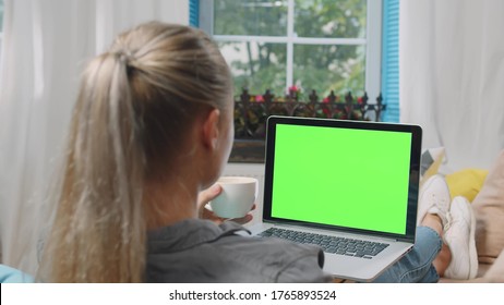 Young Woman Relaxing At Home On The Couch Having Coffee And Using Laptop Computer. Back View Of Girl Watching Movie On Laptop At Home. Green Screen